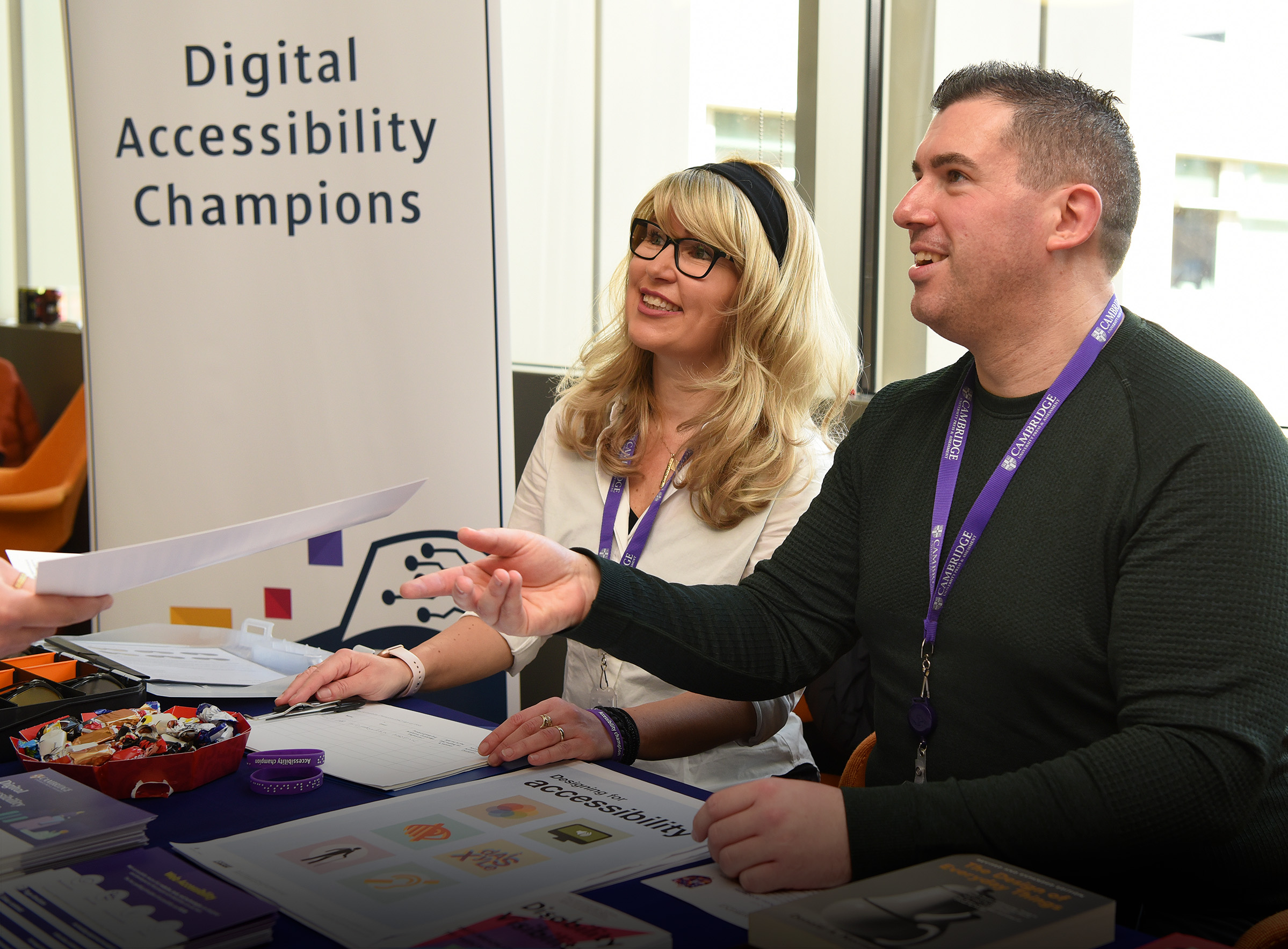 Digital Accessibility Champions, James & Julie sit side by side at their exhibition stall handing a leaflet about Digital Accessibility to someone at Digital & Data Day