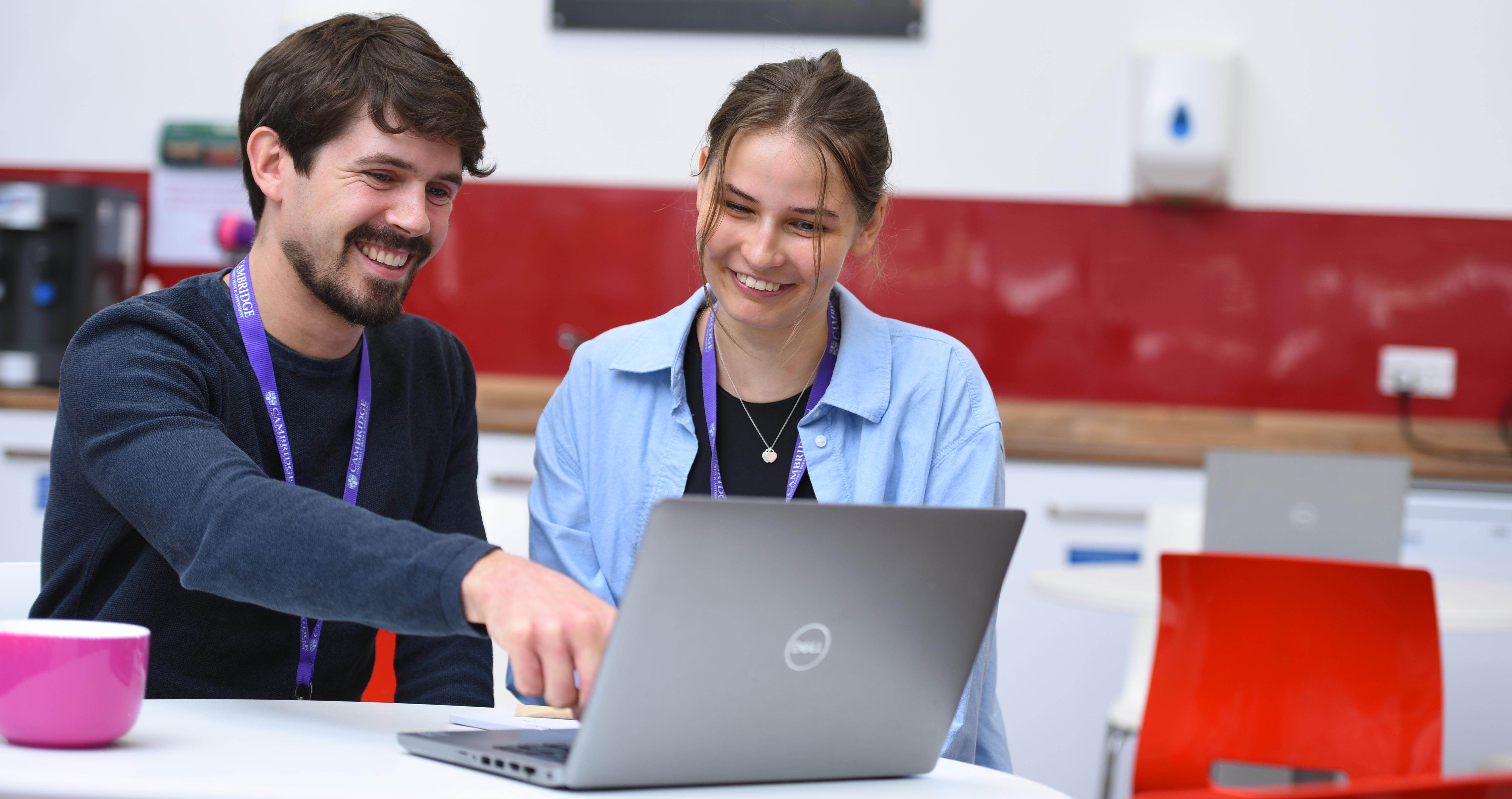 Sat in an office work setting, male colleague points toward a laptop screen next to a female colleague looking at the laptop