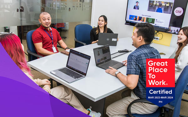 Manila colleagues sit in chairs around a desk during a meeting discussing work in front of laptops smiling with GPTW certification logo at bottom right