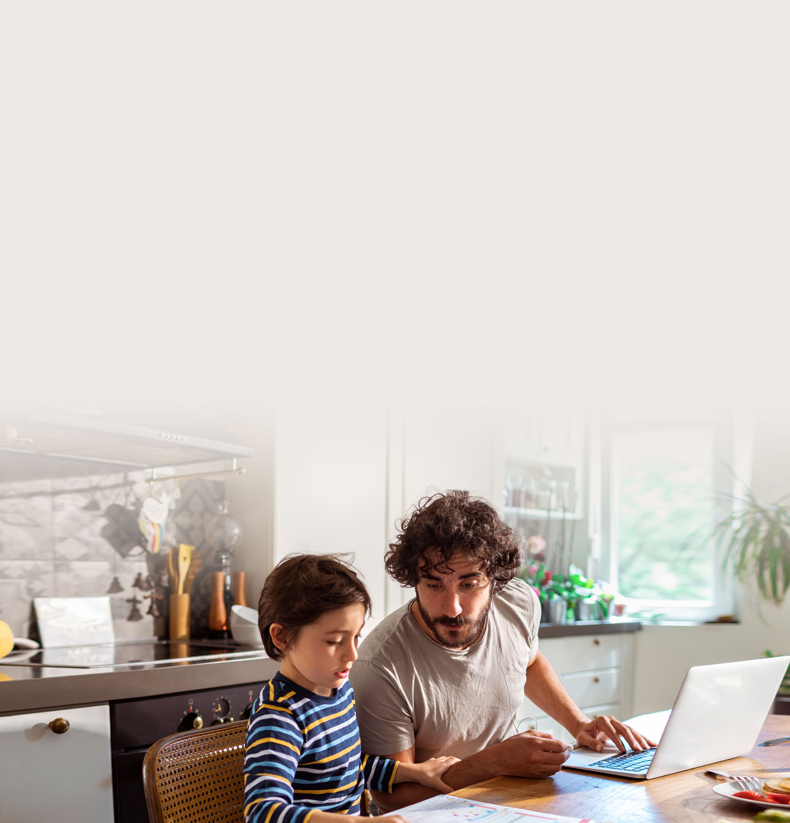 a young boy and his parent sat at a dining table with a laptop and looking at school work